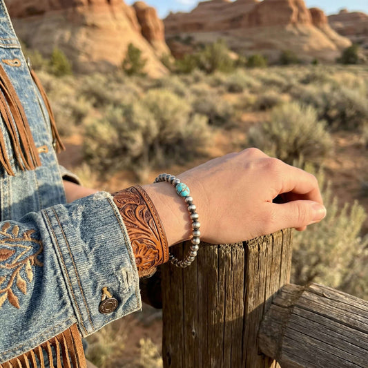 Person wearing a turquoise and silver bracelet, holding onto a wooden post in a desert landscape.