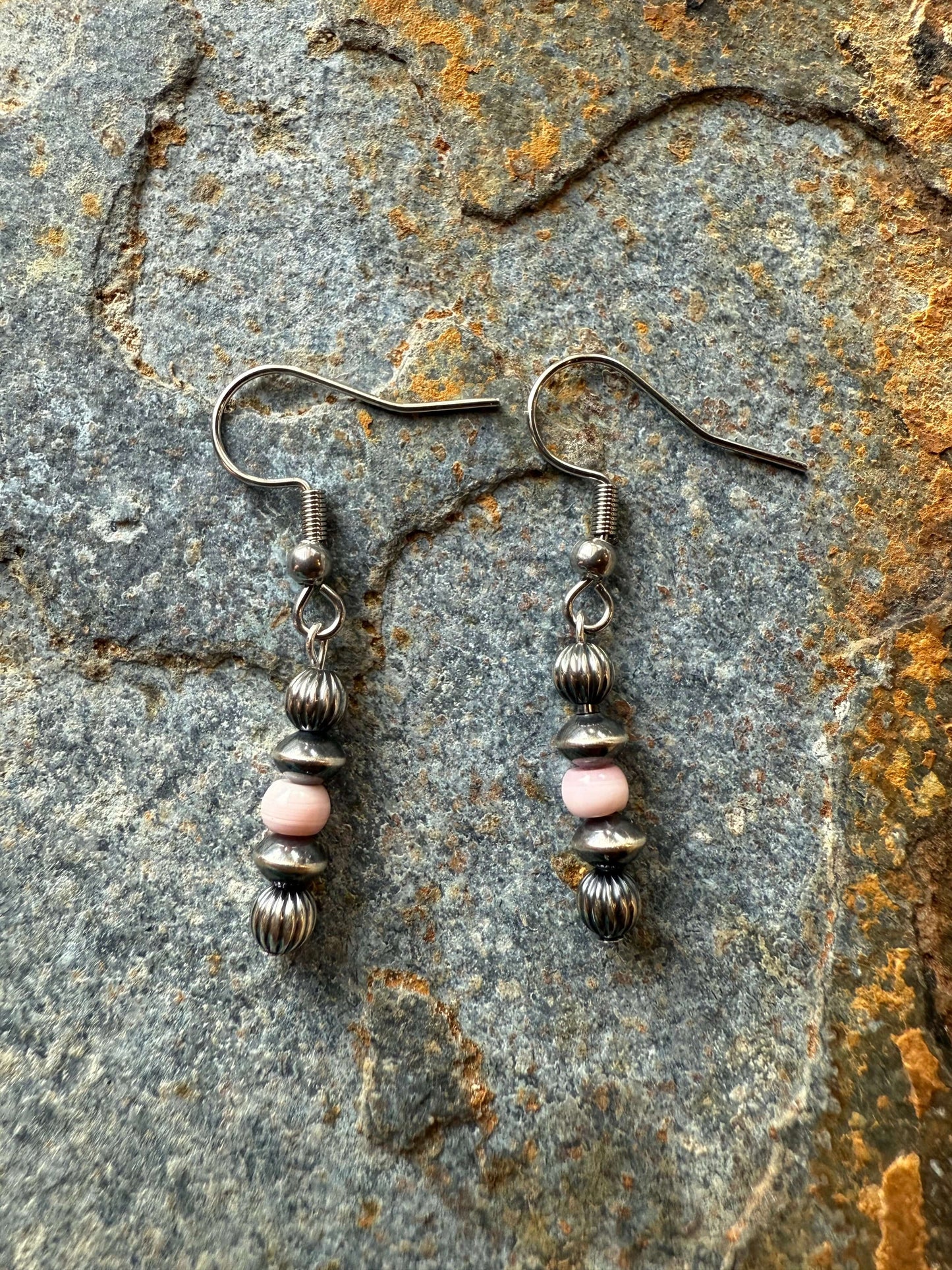 Sterling Silver Pearls & Pink Conch Shell Earrings dangle on a textured stone background, showcasing their elegance and style.
