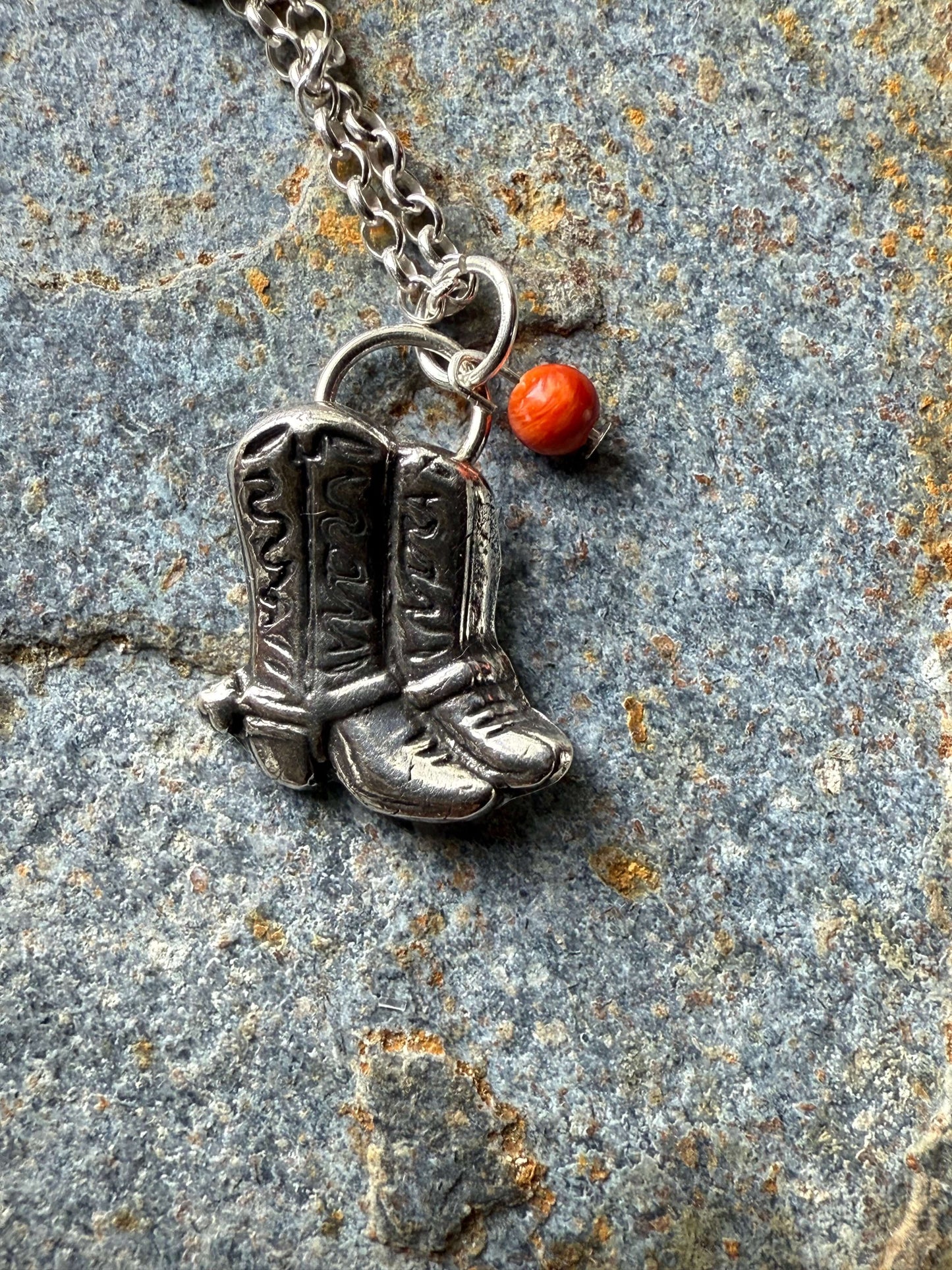 Silver cowboy boot necklace on a stone surface with red bead