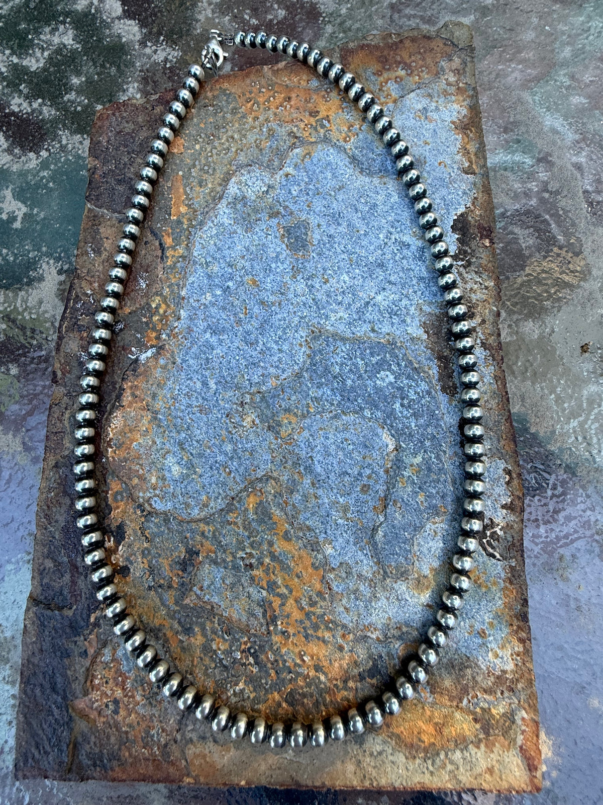 A silver beaded necklace arranged in an oval shape on a textured, rusted metal plate over a mottled gray stone surface.