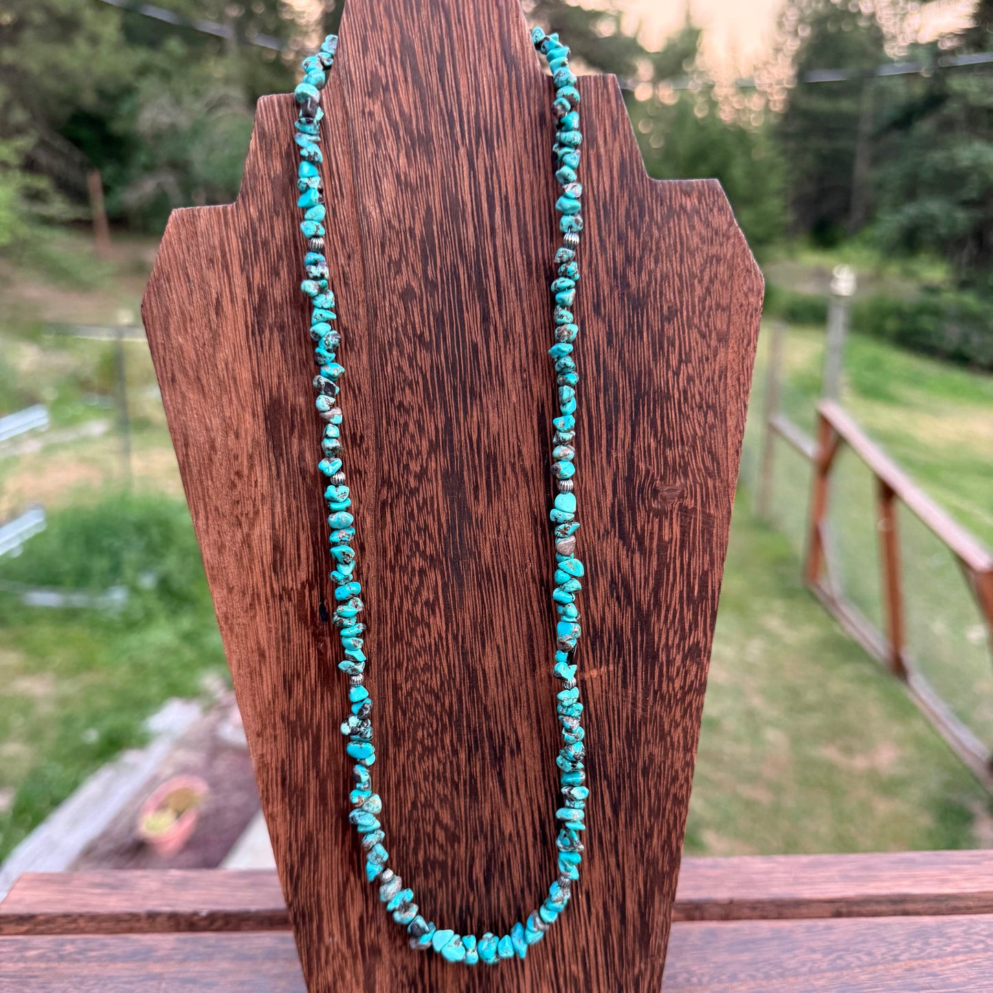 A turquoise beaded necklace draped over a dark brown wooden display stand, set against an outdoor background of green grass and trees.