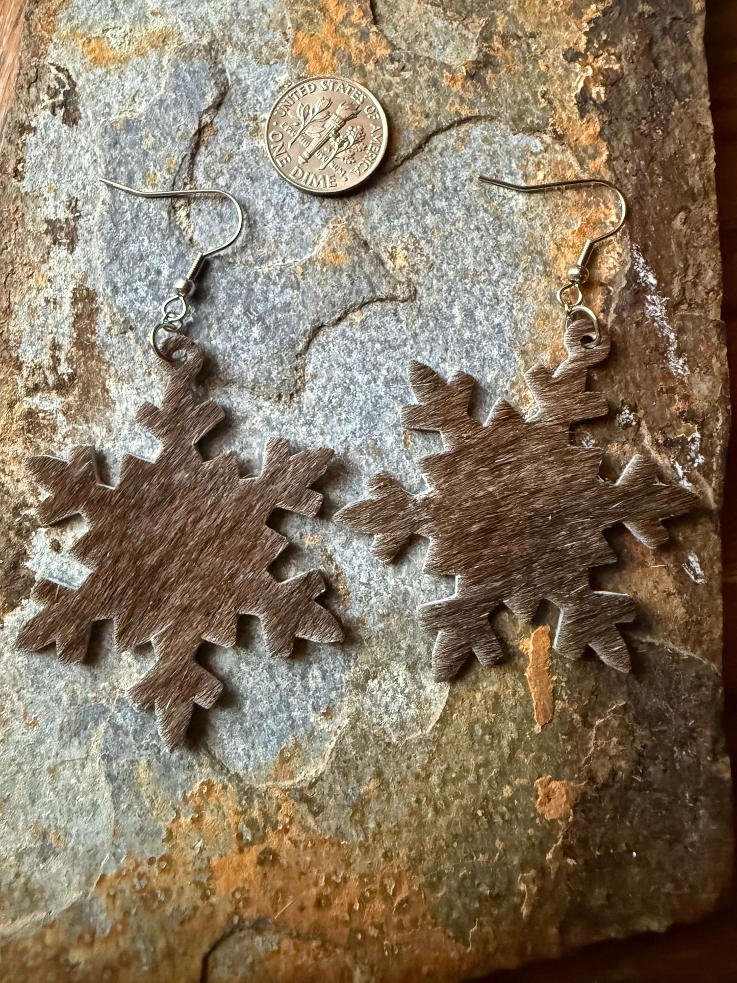Cowhide Snowflake Earrings hanging against a rustic stone background with a coin for size reference.