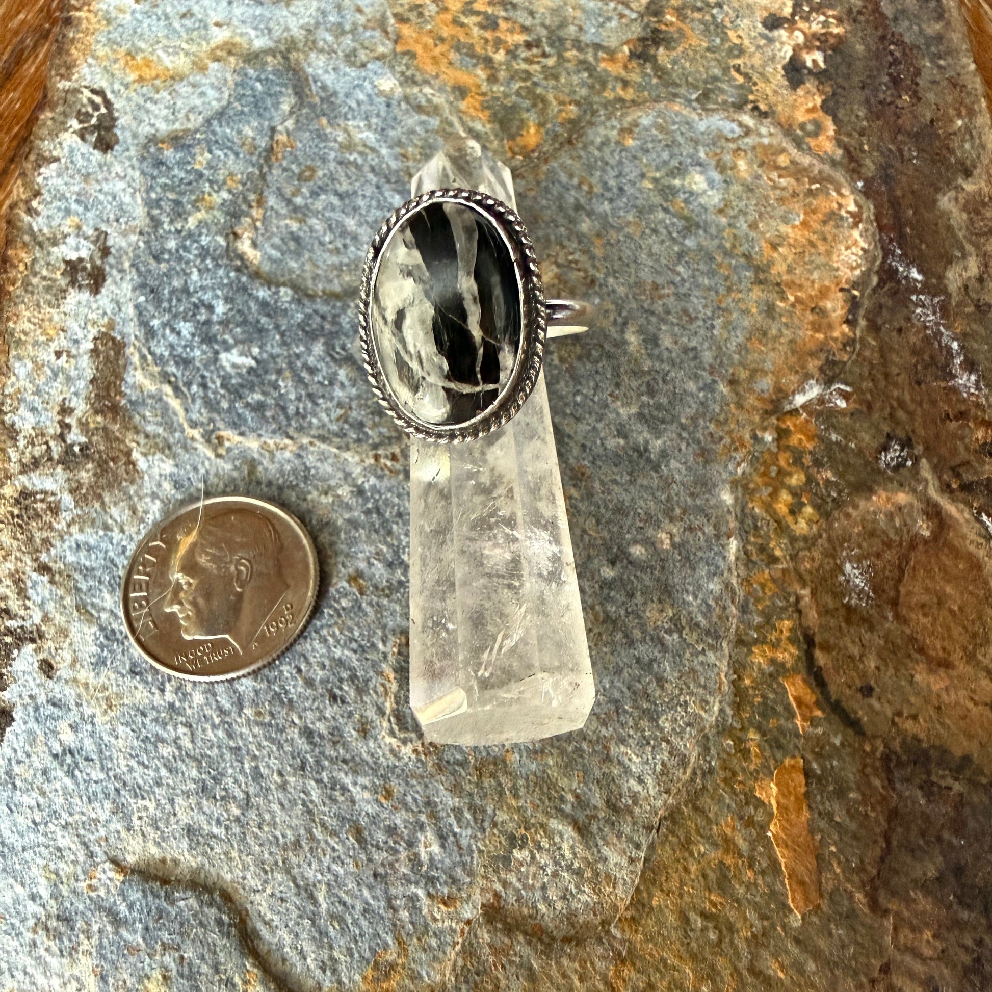 Silver ring with a black and white stone on a rock next to a coin for scale 