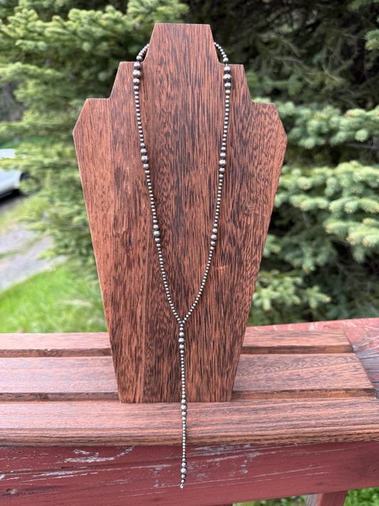 A silver beaded necklace draped over a dark brown wooden display stand, photographed outdoors with blurred green foliage and a wooden railing in the background.