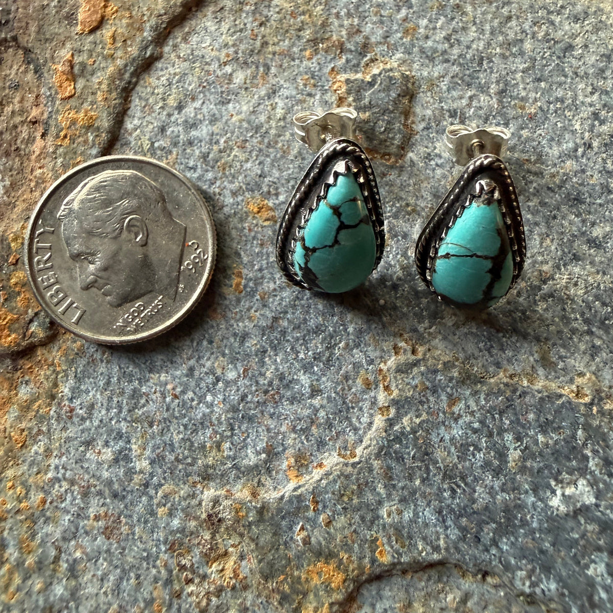 A pair of turquoise drop earrings with silver settings, displayed next to a US dime on a textured gray stone surface.