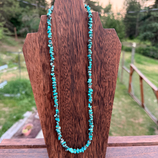 A turquoise beaded necklace draped over a dark brown wooden display stand, set against an outdoor background of green grass and trees.
