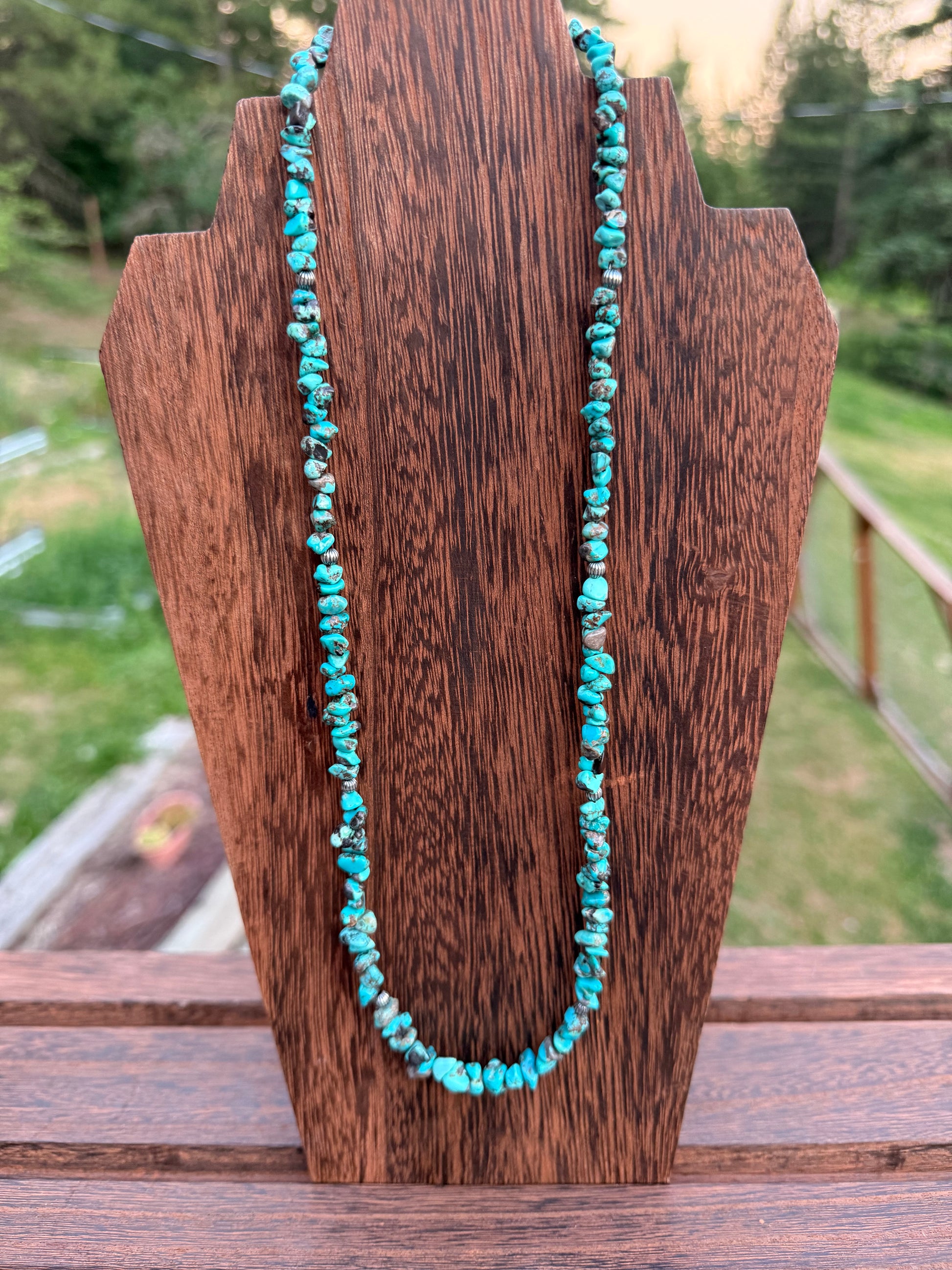 A turquoise bead necklace draped over a rustic wooden display block, with an outdoor blurred background visible behind it.