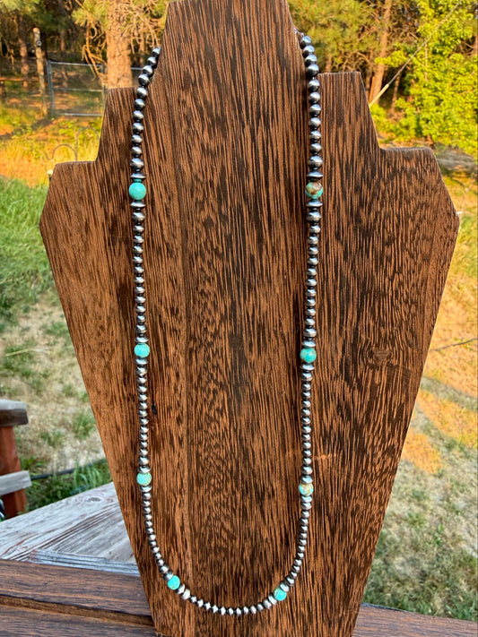 A beaded necklace with silver-toned round beads and turquoise-colored spacer beads, draped over a rustic wooden display stand with an outdoor background visible.