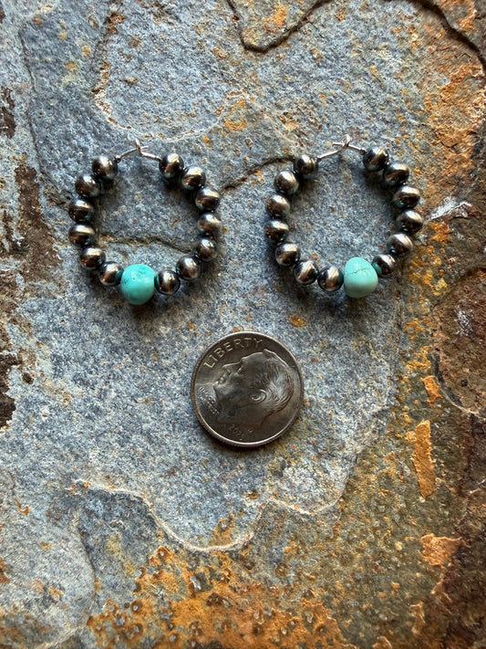 A pair of handmade hoop earrings made of dark silver beads and a light turquoise stone, displayed on a textured gray stone surface next to a US dime for scale.