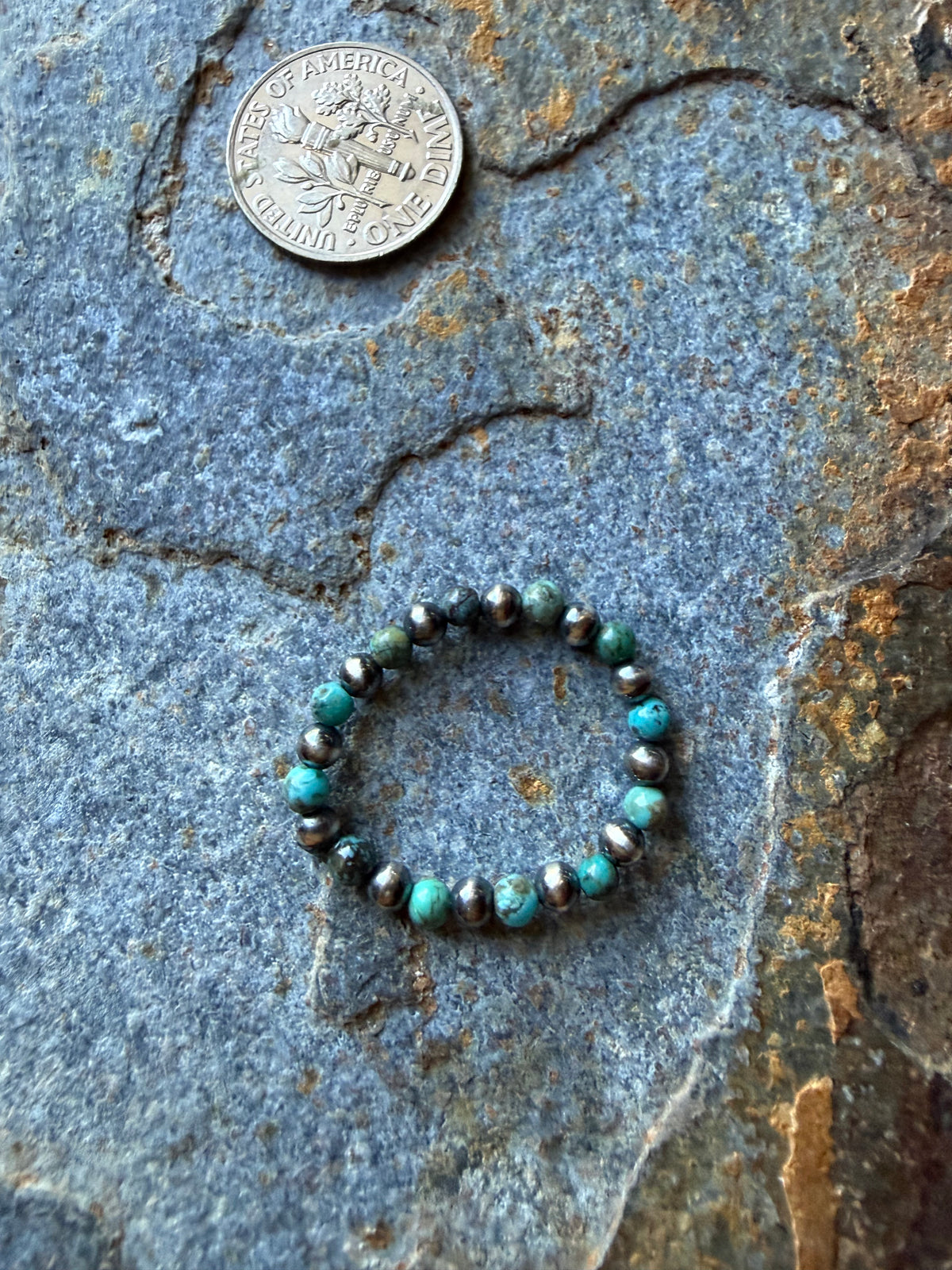 A beaded stretch ring made of turquoise, black, and metallic silver beads, displayed on a textured gray stone surface next to a US dime coin for scale.