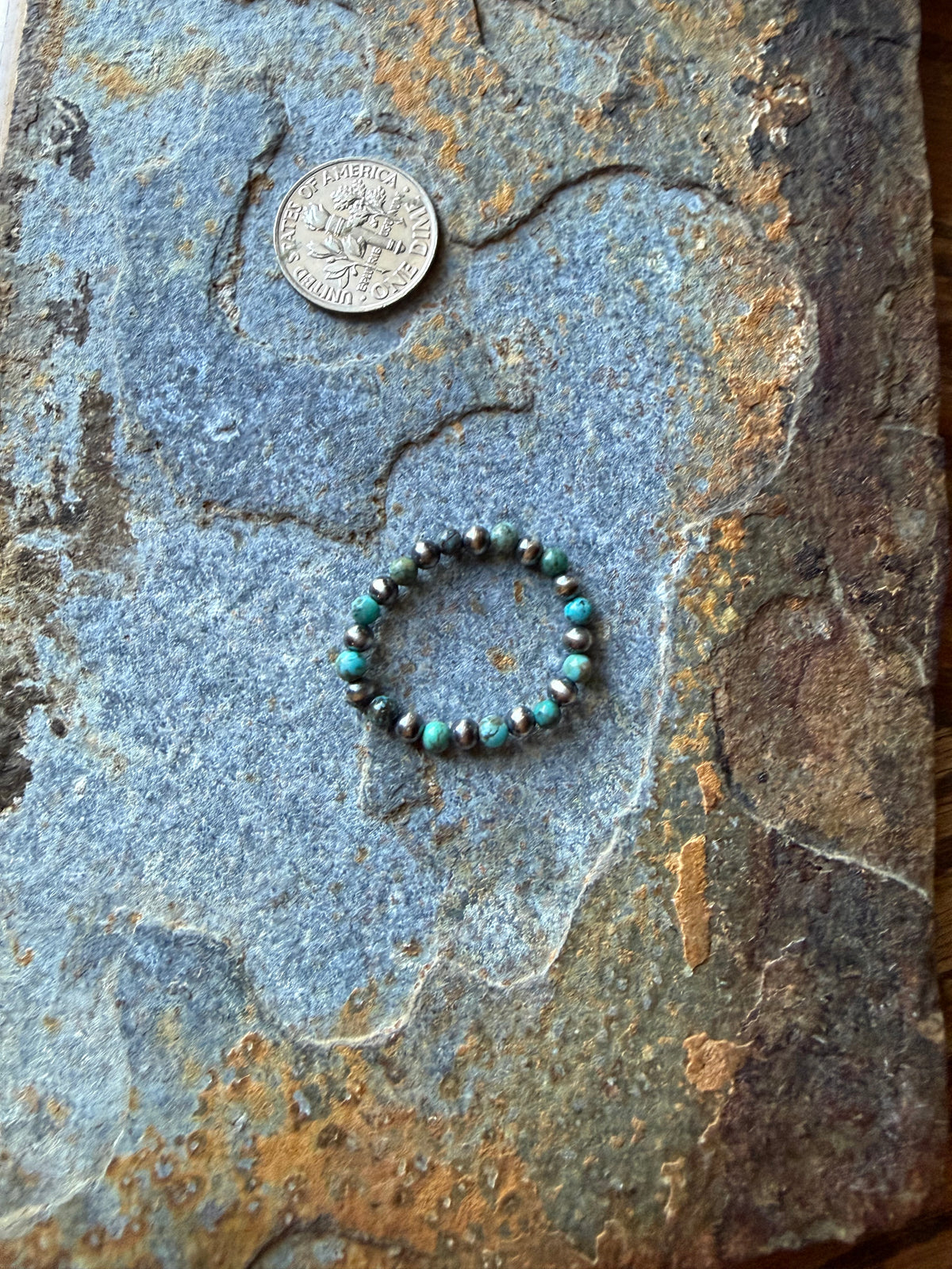 A beaded stretch ring with turquoise, black, and silver beads, displayed on a textured gray stone surface next to a US dime coin for scale.